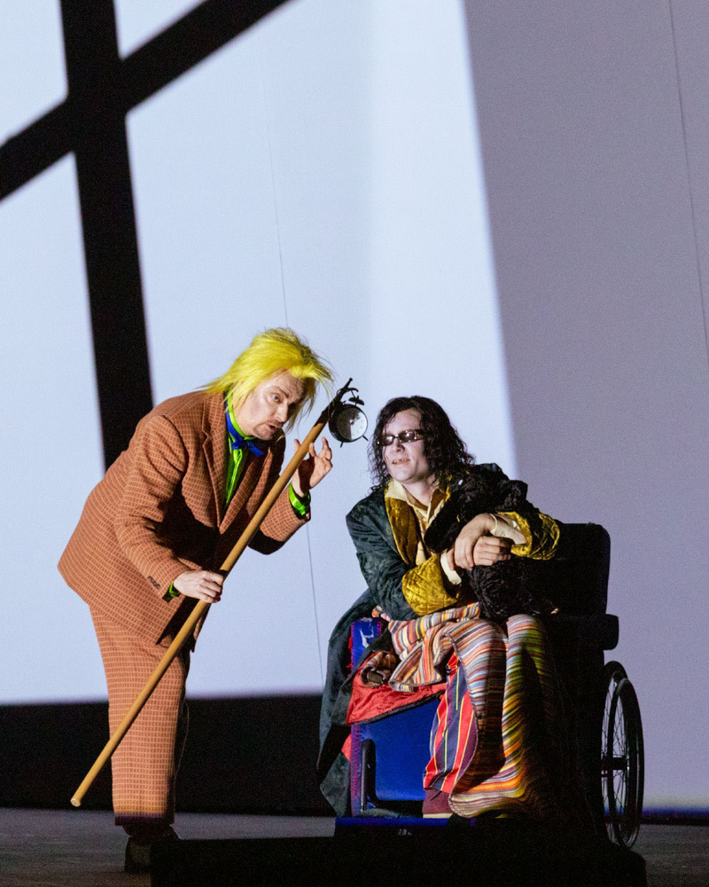 Eine Figur mit gelber Perücke beugt sich zu einer Person im Rollstuhl, beide vor einer Wand mit markanten Schattenprojektionen.