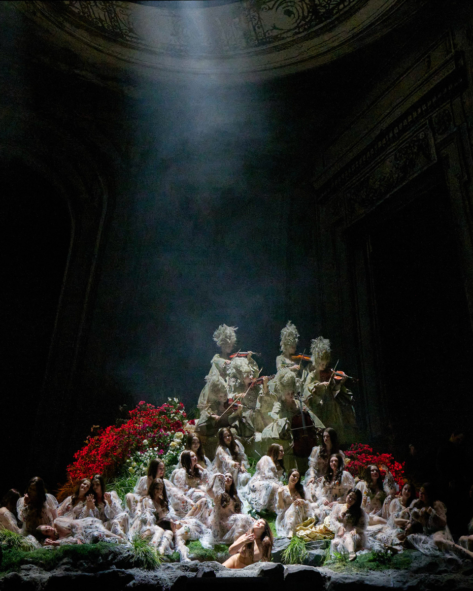 In geheimnisvollem Bühnenlicht sitzt eine Frauen-Gruppe auf einem blumenumrahmten Podest