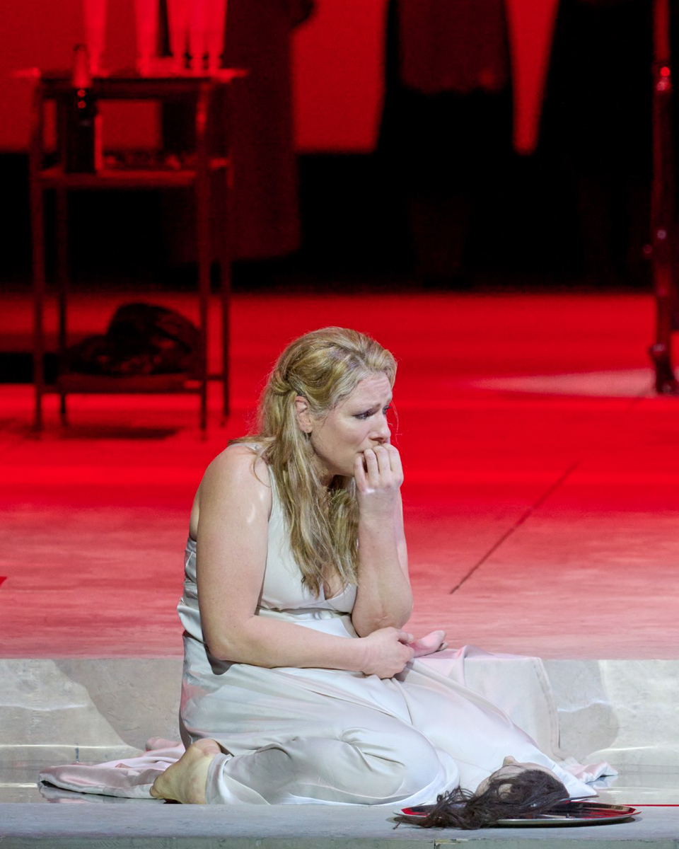 Eine Frau im weißen Kleid sitzt verzweifelt auf dem Boden einer rot ausgeleuchteten Bühne, umgeben von medizinischem Besteck und im Hintergrund stehenden Personen.