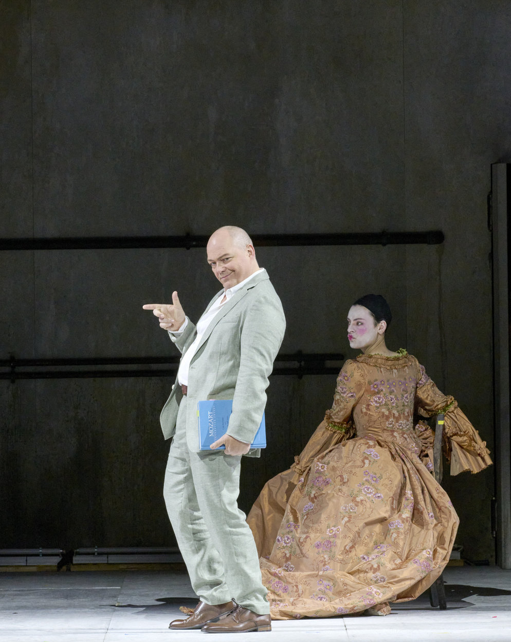 Ein lachender Mann in einem hellen Anzug zeigt mit Fingergesten nach vorne, während hinter ihm eine Frau in barocker Robe mit weiß geschminktem Gesicht auf einem Stuhl sitzt.