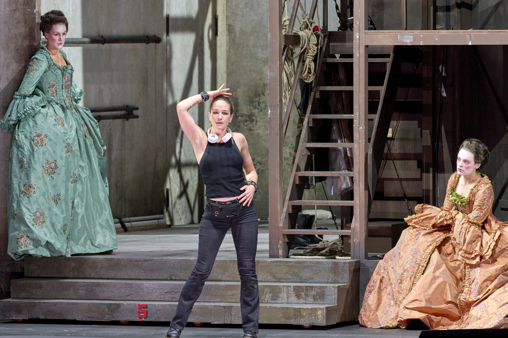 Eine Frau in schwarzem Top und dunkler Hose steht selbstbewusst mit erhobenem Arm auf der Bühne.