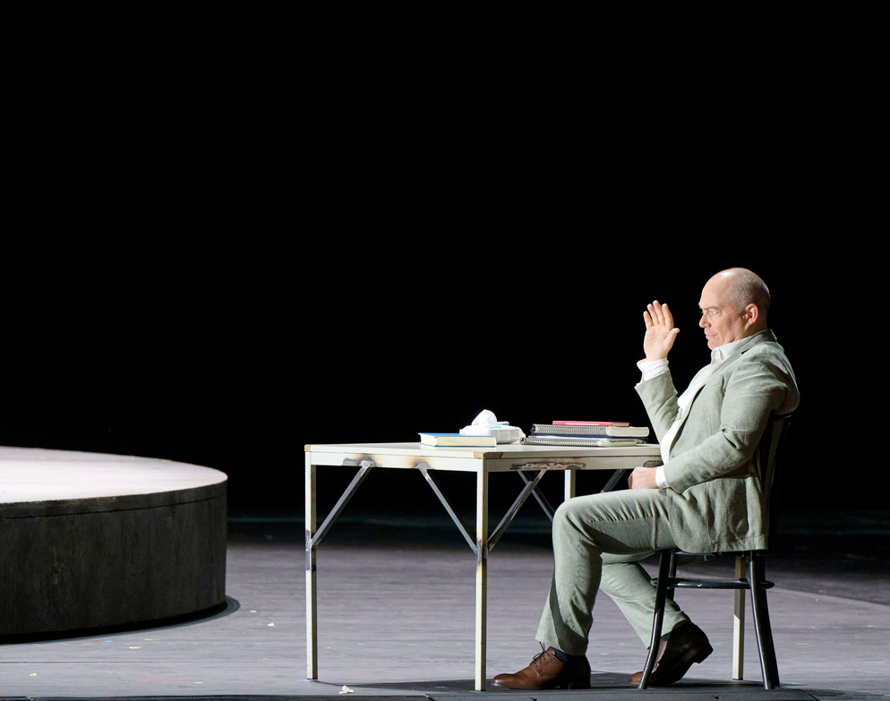 Ein Mann in hellem Anzug sitzt an einem Tisch auf einer Bühne und hebt lächelnd die Hand, als würde er winken; links ist eine runde, erhöhte Spielfläche zu sehen.