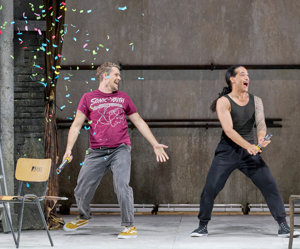 Zwei Personen lachen ausgelassen auf der Bühne, während bunte Konfettistreifen durch die Luft fliegen; eine von ihnen trägt ein T-Shirt, die andere ein ärmelloses Oberteil.